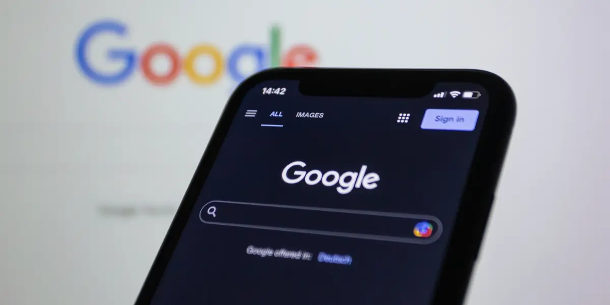 A phone showing the Google app held in front of a monitor displaying the same search page